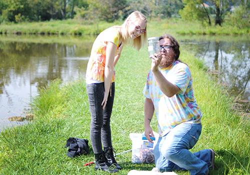 Student and professor kneel on the grass as they view water collected from a natural water source